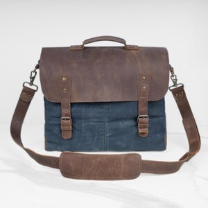 May include: A brown leather and blue canvas messenger bag with a top handle and adjustable shoulder strap. The bag features a flap closure with two buckle straps and metal hardware. The shoulder strap has a padded shoulder rest.