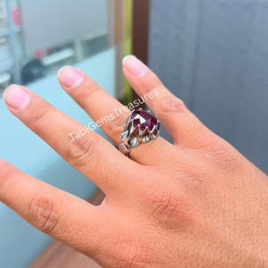 Peut inclure: Une bague en argent ornée d'une grosse pierre précieuse rouge foncé à facettes. La bague a un sertissage orné avec un motif texturé. La bague est sur une main. Le texte "TabiGemsTreasures" est visible.