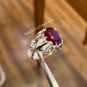 May include: A silver ring with an oval, deep red gemstone. The ring has an ornate, openwork design around the gem. The text "TabiGemsTreasures" is visible in the image.