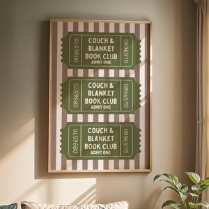 以下が含まれることがあります： ストライプの背景に「COUCH & BLANKET BOOK CLUB ADMIT ONE」の文字が入った、緑と白のチケットが3枚描かれた額入りのプリント。緑とベージュのクッションが置かれたソファの上に飾られており、背景には植物と窓が見える。