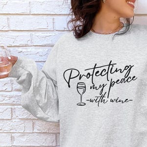 May include: A light gray sweatshirt with the text "Protecting my peace - with wine" in black script. The design includes a wine glass graphic. The sweatshirt has long sleeves and a crew neck.