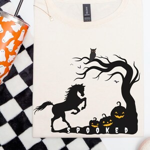 May include: Cream-colored t-shirt with a black Halloween-themed graphic featuring a horse, tree, pumpkins, and the word "SPOOKED". A tumbler with ghost illustrations and a black and white checkered pattern are also visible.