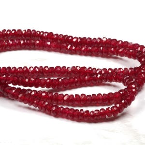 May include: Strands of faceted, deep red beads. The beads are strung together, forming a continuous loop. The beads have a sparkling, polished appearance, and are arranged against a white background.
