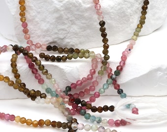 May include: Strands of colorful gemstone beads in shades of pink, brown, green, and yellow. The beads are small and round, strung together to create necklaces or bracelets. The background is a white, textured surface.