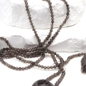 May include: Strands of faceted, smoky quartz beads. The beads are a dark gray color and are strung together. The strands are draped over a white, textured surface. Tassels are visible at the end of some strands.