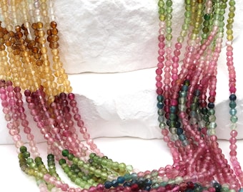 May include: Strands of colorful gemstone beads in shades of pink, green, yellow, and brown. The beads are small and round, strung together to create necklaces or bracelets. The gemstones are displayed on a white surface.