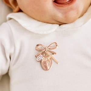 Op de afbeelding: Een baby in een witte romper met kraag en een religieuze speld in roségoud. De speld heeft een strik, een kruis, een medaillon en een kleine bedel. De baby lacht met open mond.