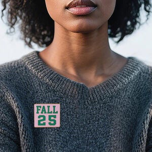 Op de afbeelding: Een roze en groene emaille pin met de tekst "FALL 25" is vastgemaakt aan een grijze trui. De pin is vierkant en de tekst is in een vetgedrukte, schreefloze lettertype. De trui heeft een V-hals.