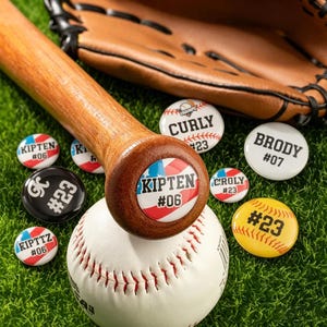 Puede incluir: Un bate de béisbol de madera descansa sobre una pelota de béisbol, rodeado de botones personalizados con temática de béisbol. Los botones muestran nombres y números, con un guante de béisbol en el fondo. El extremo del bate tiene un diseño rojo, blanco y azul.