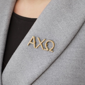 May include: A gold-coloured brooch displaying the Greek letters AXΩ, embellished with small, round, white pearls. The brooch is attached to a grey blazer, with a black top visible underneath.