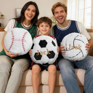 Op de afbeelding: Drie pluche sportballen: een honkbal met "Annabette" in roze, een voetbal met "Jack" in goud en een volleybal met "Jack" in goud. De ballen zijn wit met zwarte en rode accenten.