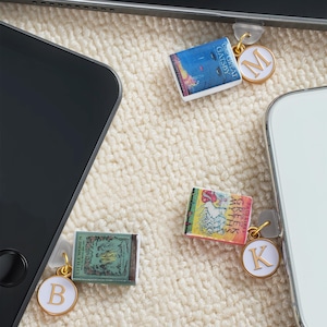 Puede incluir: Miniaturas de libros con detalles dorados y colgantes iniciales de esmalte blanco. Los libros incluyen títulos como "The Great Gatsby", "Greek Myths" y "Little Women". Los colgantes están unidos a teléfonos inteligentes.