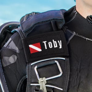 Op de afbeelding: Een zwarte stoffen patch bevestigd aan de schouderband van een duiktrimvest. De rechthoekige patch toont links het rood-witte duikvlag-symbool en rechts de witte tekst Toby op een zwarte achtergrond.