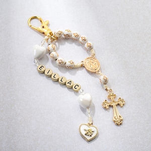 Puede incluir: Un llavero de rosario blanco y dorado con un cierre dorado. El rosario presenta cuentas blancas con cruces doradas, un colgante de cruz dorada y un dije de corazón dorado con un diseño de ángel. El nombre "EILLAC" está escrito con letras doradas.