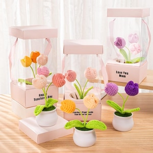 May include: Crocheted tulip flowers in white pots, displayed in pink boxes with clear sides. Some boxes have text that reads "Happy Teacher's Day" and "Love You Mom". The flowers are various colors, including yellow, orange, pink, and purple.