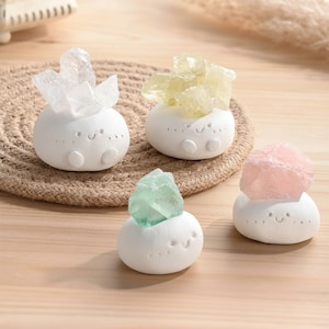 May include: Four white ceramic figurines, each topped with a different colored crystal: clear, yellow, green, and pink. The figurines have a rounded shape with a smiling face and small feet. They are arranged on a wooden surface.