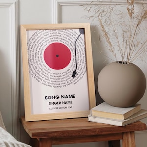Op de afbeelding: Een ingelijste print met een rood vinylplaatontwerp en songteksten in een cirkelvormig patroon. De print heeft een licht houten frame en ruimte voor songdetails. Een vaas en boeken staan op een houten tafel. De print is 30 cm x 40 cm groot.