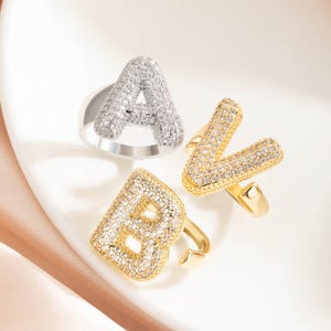 May include: Three letter rings, featuring the letters A, B, and V. The rings are encrusted with small, sparkling stones. The ring for the letter A is silver, while the rings for the letters B and V are gold.