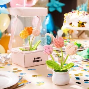 Może przedstawiać: Różowe pudełko prezentowe z przezroczystą pokrywą zawiera szydełkowe kwiaty w odcieniach żółci, pomarańczy i różu. Na pudełku widnieje napis "Happy birthday". Biała doniczka z szydełkowymi kwiatami stoi przed pudełkiem.