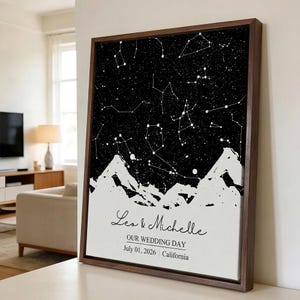 Puede incluir: Una impresión artística enmarcada con un diseño celestial en blanco y negro. La obra de arte representa constelaciones sobre una cordillera. Los nombres "Lea & Michelle", "OUR WEDDING DAY", la fecha y el lugar están impresos en la parte inferior.