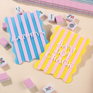 Pode incluir: Duas bases decorativas com bordas onduladas e listras verticais. Uma é azul e rosa com o nome "KATHRYN", a outra é amarela e rosa com "BAM DOT CRACK". Peças de mahjong espalhadas.