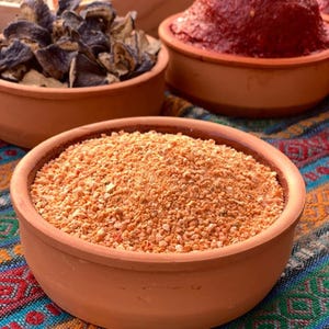 Puede incluir: Primer plano de varios alimentos en cuencos de terracota. El primer plano presenta un cuenco lleno de comida granular de color marrón claro. Otros cuencos contienen verduras secas de color púrpura oscuro y una pasta roja. Los cuencos están sobre un textil estampado colorido.