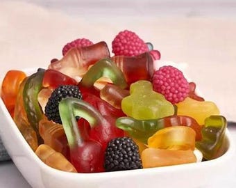 May include: A white square bowl filled with a colorful assortment of gummy candies. The candies include red cherries, green bears, and black blackberries. The bowl is set against a soft, neutral background.