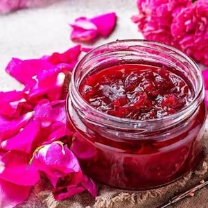 May include: A glass jar filled with red jam, showing pieces of fruit, is placed on a hessian cloth. Bright pink flower petals and blooms are scattered around the jar, with a small spoon beside it. The image suggests homemade preserves.