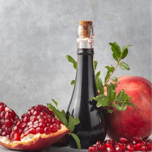 Puede incluir: Una botella de vidrio oscuro con tapón de corcho, llena de líquido oscuro, junto a una granada cortada por la mitad y una granada entera. Hojas verdes y semillas de granada sueltas están esparcidas alrededor de la botella y la fruta.