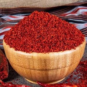 May include: A wooden bowl filled with vibrant red chili flakes, with a pile of flakes and dried red chilies scattered around. The bowl sits on a dark surface, with a patterned fabric in the background. Culinary and food preparation.