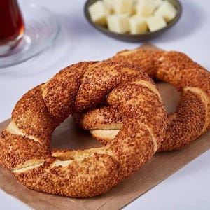 Könnte beinhalten: Zwei mit Sesam bedeckte Bagels liegen auf braunem Papier. Im Hintergrund befinden sich eine kleine Schüssel mit gewürfeltem Käse und ein Glas Tee. Die Bagels sind goldbraun und sehen frisch gebacken aus.