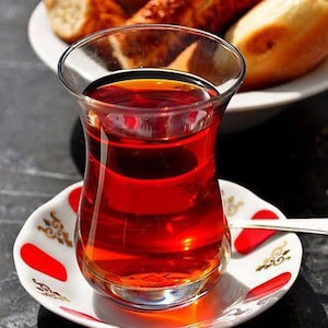 May include: A glass of Turkish tea, filled with a rich, red liquid, sits on a decorative saucer with red accents. A plate of bread is in the background. The tea glass has a unique, tulip-like shape.