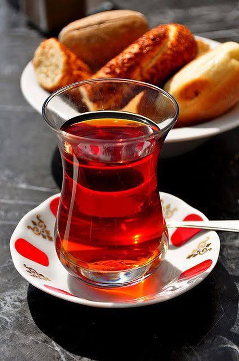 May include: A glass of Turkish tea, filled with a rich, red liquid, sits on a decorative saucer with red accents. A plate of bread is in the background. The tea glass has a unique, tulip-like shape.