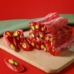 K&ouml;nnte beinhalten: Nahaufnahme von t&uuml;rkischem Honig, einer traditionellen S&uuml;&szlig;ware. Die leuchtend rote S&uuml;&szlig;igkeit ist mit gr&uuml;nen Pistazien &uuml;bers&auml;t und auf einem Holzschneidebrett vor rotem Hintergrund angeordnet. Die S&uuml;&szlig;igkeit ist in rechteckige Formen geschnitten.
