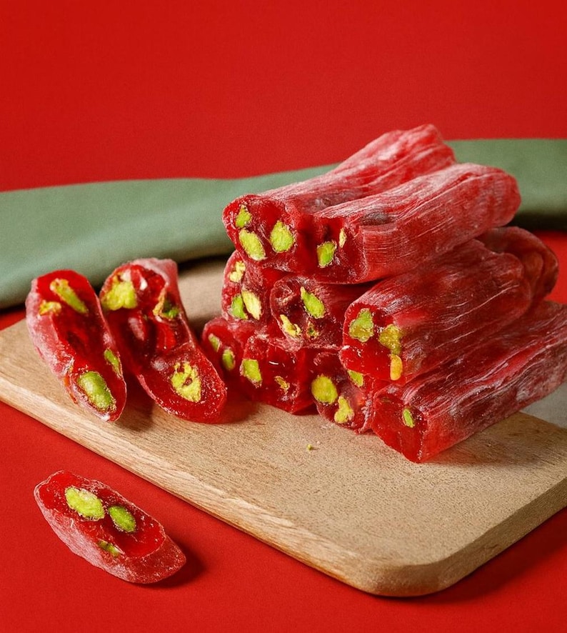 K&ouml;nnte beinhalten: Nahaufnahme von t&uuml;rkischem Honig, einer traditionellen S&uuml;&szlig;ware. Die leuchtend rote S&uuml;&szlig;igkeit ist mit gr&uuml;nen Pistazien &uuml;bers&auml;t und auf einem Holzschneidebrett vor rotem Hintergrund angeordnet. Die S&uuml;&szlig;igkeit ist in rechteckige Formen geschnitten.