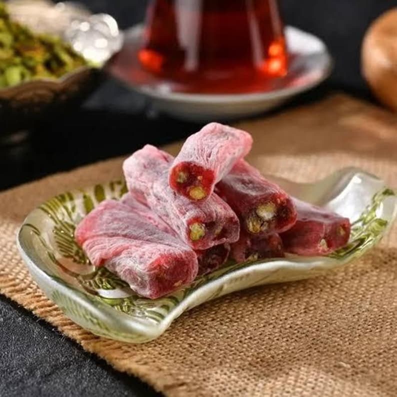 K&ouml;nnte beinhalten: Nahaufnahme von t&uuml;rkischem Honig, einer traditionellen S&uuml;&szlig;ware, auf einem dekorativen Teller. Die rosa, rechteckigen S&uuml;&szlig;igkeiten sind mit Puderzucker best&auml;ubt und mit Pistazien gef&uuml;llt. Eine Tasse Tee und eine Sch&uuml;ssel Pistazien befinden sich im Hintergrund.