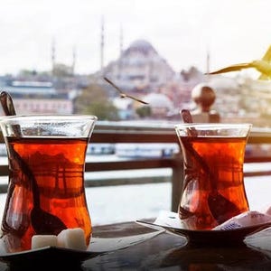 May include: Two glasses of tea, each with a spoon, on small saucers, with sugar cubes. A city with a mosque and seagulls in flight forms the background. The tea is a deep amber colour.