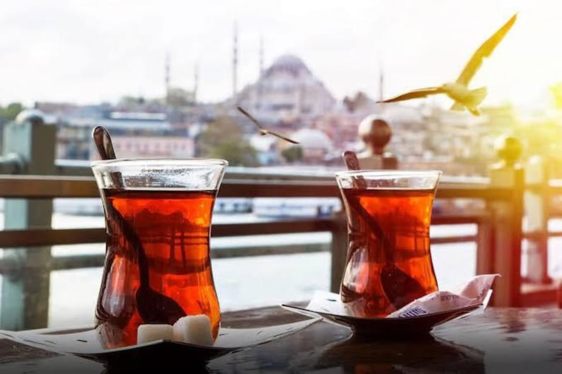 May include: Two glasses of tea, each with a spoon, on small saucers, with sugar cubes. A city with a mosque and seagulls in flight forms the background. The tea is a deep amber colour.
