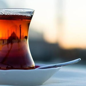 May include: A traditional glass of tea, filled with amber liquid, rests on a white saucer with a spoon. The glass has a distinctive hourglass shape. The tea reflects a blurred cityscape, suggesting a warm drink.