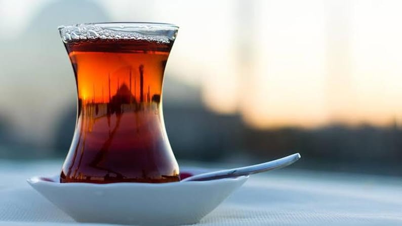 May include: A traditional glass of tea, filled with amber liquid, rests on a white saucer with a spoon. The glass has a distinctive hourglass shape. The tea reflects a blurred cityscape, suggesting a warm drink.