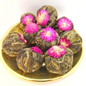 May include: A collection of tea balls, each wrapped in dried leaves and topped with a vibrant pink flower, presented on a gold-colored plate. The tea balls are a light green color, and the flowers are a bright pink.