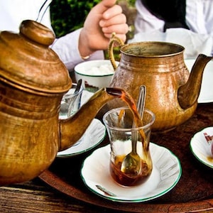 May include: A copper teapot pours tea into a clear glass with a spoon, on a wooden tray. Two other copper teapots and two glasses of tea are also on the tray. The tea is served with small white saucers and a small bowl of sugar.