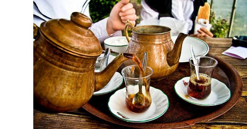 May include: A copper teapot pours tea into a clear glass with a spoon, on a wooden tray. Two other copper teapots and two glasses of tea are also on the tray. The tea is served with small white saucers and a small bowl of sugar.