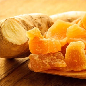 May include: Close-up of fresh ginger root and candied ginger pieces. The ginger root is light brown with a tan interior. The candied ginger is a bright orange color and sits on a wooden spoon. The background is a warm, wooden surface.