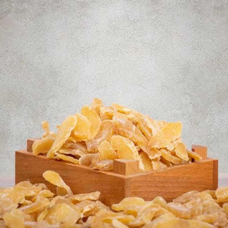 May include: A wooden box overflowing with candied ginger slices. The ginger is a light yellow color and appears sugared. The box is made of light brown wood and sits on a white surface against a gray background.