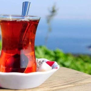 May include: A glass of Turkish tea, filled with a rich, red liquid, sits on a white saucer with a colourful rim. A small spoon rests in the tea. The background features a blurred view of the sea and greenery.