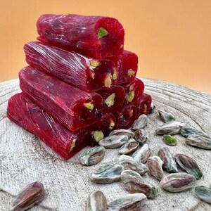 K&ouml;nnte beinhalten: Ein Stapel leuchtend roter t&uuml;rkischer K&ouml;stlichkeiten, besetzt mit gr&uuml;nen Pistazien. Die S&uuml;&szlig;waren sind auf einer Holzoberfl&auml;che angeordnet, mit verstreuten Pistazien in der N&auml;he. Der Hintergrund ist ein warmes Orange.