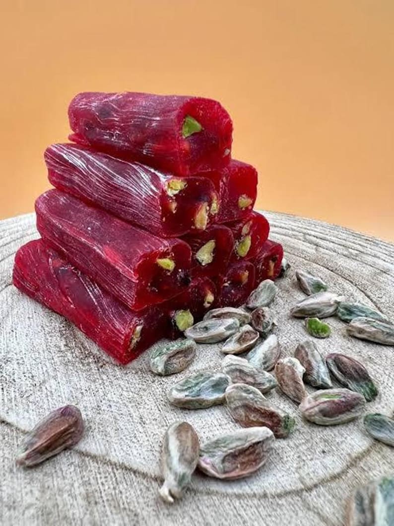 K&ouml;nnte beinhalten: Ein Stapel leuchtend roter t&uuml;rkischer K&ouml;stlichkeiten, besetzt mit gr&uuml;nen Pistazien. Die S&uuml;&szlig;waren sind auf einer Holzoberfl&auml;che angeordnet, mit verstreuten Pistazien in der N&auml;he. Der Hintergrund ist ein warmes Orange.