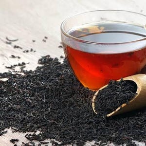 May include: A glass teacup filled with dark tea sits on a wooden surface, surrounded by loose black tea leaves. A wooden scoop holds more tea leaves. The tea has a rich, amber colour.
