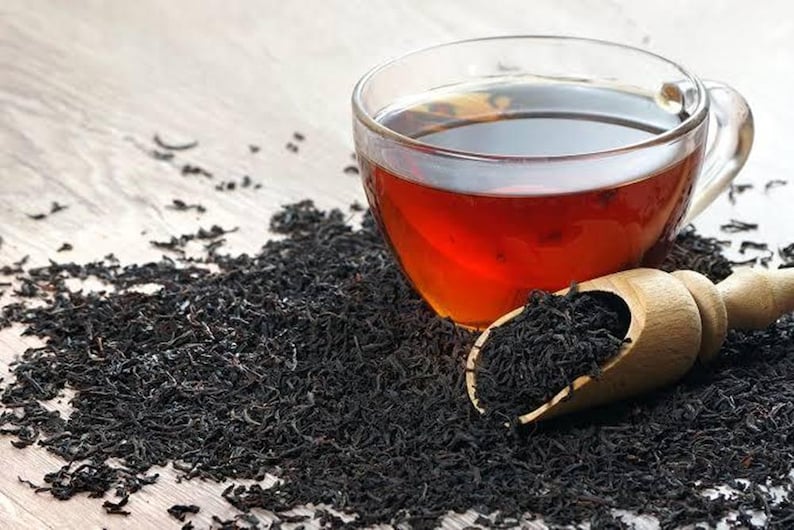 May include: A glass teacup filled with dark tea sits on a wooden surface, surrounded by loose black tea leaves. A wooden scoop holds more tea leaves. The tea has a rich, amber colour.